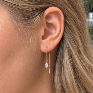 May include: A close-up of a gold-colored earring with a dangling, marquise-cut, clear gemstone. The earring is a threader style, with a delicate chain that goes through the ear. The earring is worn on a person's ear.