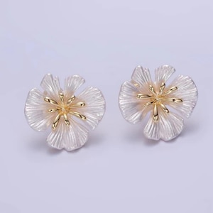 May include: A pair of flower-shaped stud earrings. Each earring features a white, textured petal design with a gold-colored center. The earrings are set against a light purple background.