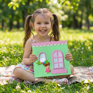 May include: A young girl smiles while holding a handmade green and pink felt house. The house features a window, a door, and a small doll. The background is a sunny outdoor scene with green grass and trees.
