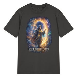May include: A charcoal grey t-shirt featuring a graphic print of a hooded figure standing before a glowing, golden circular portal. The design includes swirling cosmic light effects and the text I STEP THROUGH A PORTAL INTO A NEW VERSION OF REALITY.