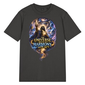 Op de afbeelding: Een antracietgrijs T-shirt met korte mouwen en een centrale afbeelding van een figuur omringd door wervelende kosmische nevels, planeten en sterren. De tekst "THE UNIVERSE MOVES IN HARMONY WITH ME" staat in goudkleurige en blauwe letters gedrukt.