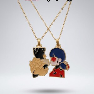 May include: A gold-toned necklace set featuring two pendants that form a pair. One pendant depicts a black and tan fox-like character, while the other shows a character with dark blue hair and a red, black-spotted suit. Text reads Reorah x Miraculous.