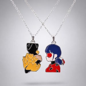 May include: Two silver-toned metal necklaces featuring enamel pendants. One pendant depicts a stylised black and yellow floral shape, while the other shows a character with dark blue hair, a red mask, and a red and black polka-dot patterned outfit.