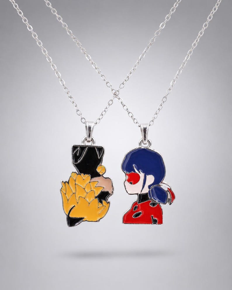 May include: Two silver-toned metal necklaces featuring enamel pendants. One pendant depicts a stylised black and yellow floral shape, while the other shows a character with dark blue hair, a red mask, and a red and black polka-dot patterned outfit.