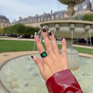 May include: A black ring featuring a vibrant green paw print design on the front face. The ring is worn on a finger with almond-shaped nails that have a nude base and dark brown floral-like tips, set against a blurred outdoor fountain.