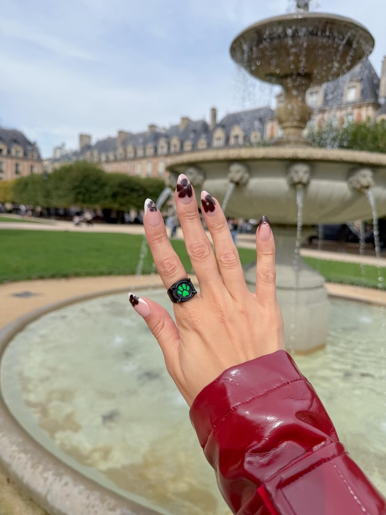 May include: A black ring featuring a vibrant green paw print design on the front face. The ring is worn on a finger with almond-shaped nails that have a nude base and dark brown floral-like tips, set against a blurred outdoor fountain.
