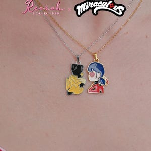 May include: Two necklaces from the Reorah Collection x Miraculous collaboration. One silver-tone chain features a black and yellow pendant, while the gold-tone chain features a pendant depicting a character with blue hair in a red and black outfit.
