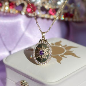 Puede incluir: Collar dorado con un colgante ovalado con un diseño de sol y una piedra preciosa púrpura central. El colgante está rodeado de pequeños cristales transparentes. El collar descansa sobre una caja de joyería blanca con un diseño de sol.