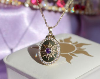 May include: Gold-coloured necklace with an oval pendant featuring a sunburst design and a central purple gemstone. The pendant is surrounded by small, clear crystals. The necklace rests on a white jewellery box with a sun design.