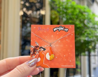 May include: A silver-toned necklace featuring a fox tail pendant with orange and white enamel details, displayed on an orange Miraculous branded card with a cartoon character illustration and the text Miraculous and 2026 Miraculous Corp.