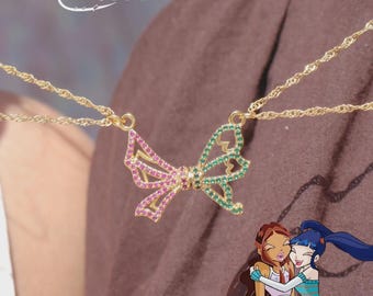May include: A gold-toned necklace featuring a bow-shaped pendant adorned with pink and green crystals. The text Winx Club x Reorah Collection is visible at the top, and an illustration of two animated characters is positioned at the bottom right.