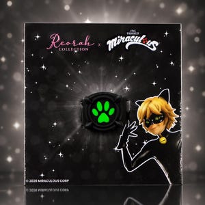 May include: A black ring featuring a glowing green paw print design, presented on a black display card with the text Reorah Collection and Miraculous, featuring an illustration of a character with blonde hair and a black mask, dated 2026.