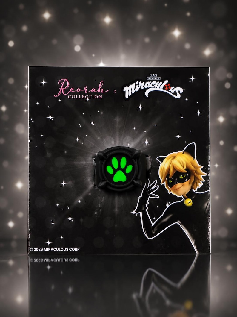 May include: A black ring featuring a glowing green paw print design, presented on a black display card with the text Reorah Collection and Miraculous, featuring an illustration of a character with blonde hair and a black mask.