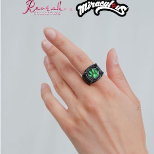 May include: A black ring featuring a central green paw print emblem. The text Reorah Collection x Miraculous is displayed above, with copyright information at the bottom. The ring has a dark, metallic finish with raised detailing around the green center.