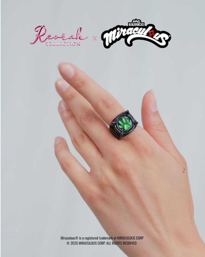 May include: A black ring featuring a central green paw print emblem. The text Reorah Collection x Miraculous is displayed above, with copyright information at the bottom. The ring has a dark, metallic finish with raised detailing around the green center.
