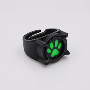 May include: A black ring featuring a circular centerpiece with a vibrant green paw print design. The ring has a matte finish and a thick, adjustable band structure with four small prongs surrounding the central paw print graphic.