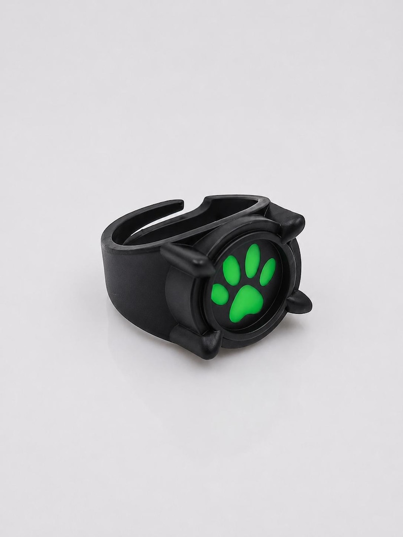 May include: A black ring featuring a circular centerpiece with a vibrant green paw print design. The ring has a matte finish and a thick, adjustable band structure with four small prongs surrounding the central paw print graphic.