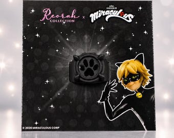 May include: A black ring featuring a central paw print design, mounted on a black display card with the text Reorah Collection and Miraculous, featuring an illustration of a character with blonde hair and a black mask. © 2026 Miraculous Corp.