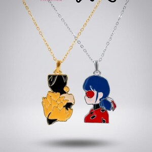 May include: Two necklaces featuring enamel pendants from the Reorah Collection x Miraculous collaboration. One gold-toned chain holds a black and yellow fox-themed pendant, while the silver-toned chain holds a red, blue, and black Ladybug character pendant.