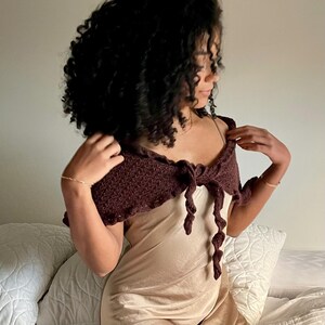 May include: A dark brown crochet shoulder wrap with a tie closure. The wrap is worn over a light tan slip dress. The crochet design is visible, and the wrap appears to be handmade. The overall aesthetic is bohemian and stylish.