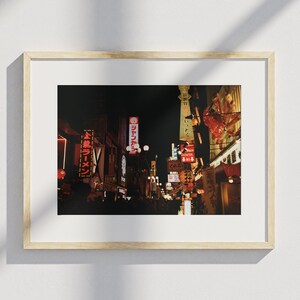 May include: A framed photograph of a busy Japanese street at night. Neon signs in Japanese characters light up the buildings, creating a warm glow on the street. The image captures the lively atmosphere of a city.
