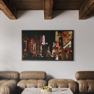 May include: A framed print depicting a Japanese street scene at night, with glowing signs and shop fronts. The image has a warm colour palette, with red, yellow, and orange tones. The print is displayed above a brown leather sofa.