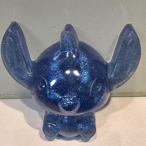 Op de afbeelding: Een blauw glitterfiguur van het Disney-personage Stitch. Het beeldje heeft grote oren, een kleine neus en een brede glimlach. Het figuur staat rechtop met de armen langs de zijden. Het beeldje is gemaakt van een glanzend, glinsterend materiaal.