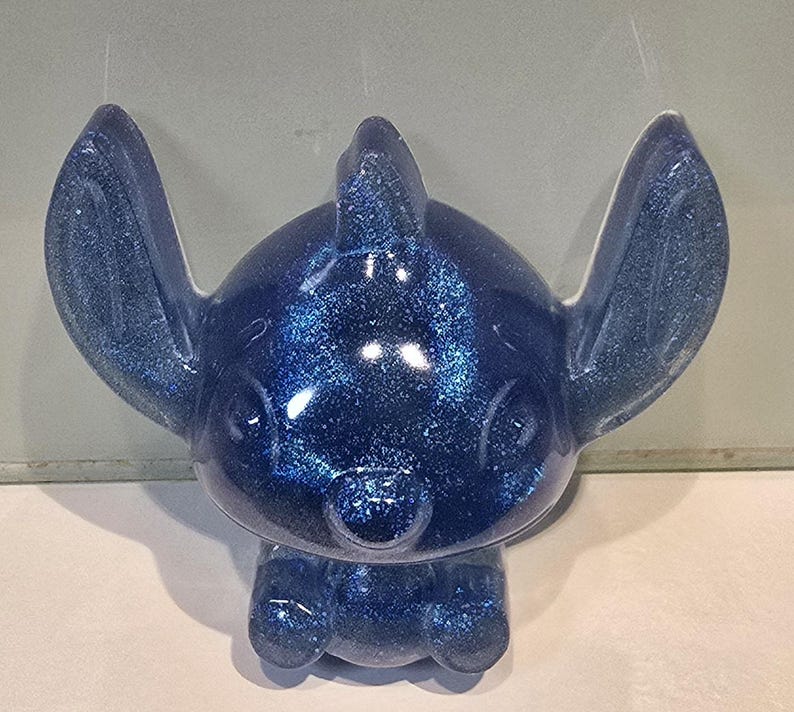 Op de afbeelding: Een blauw glitterfiguur van het Disney-personage Stitch. Het beeldje heeft grote oren, een kleine neus en een brede glimlach. Het figuur staat rechtop met de armen langs de zijden. Het beeldje is gemaakt van een glanzend, glinsterend materiaal.