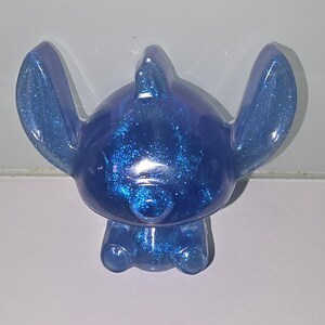 Op de afbeelding: Een blauw glitterfiguur van een stripfiguur met grote oren, een kleine neus en een afgerond lichaam. Het beeldje heeft een effen kleur met ingebedde glitter, waardoor het een sprankelend uiterlijk heeft. Het personage zit.