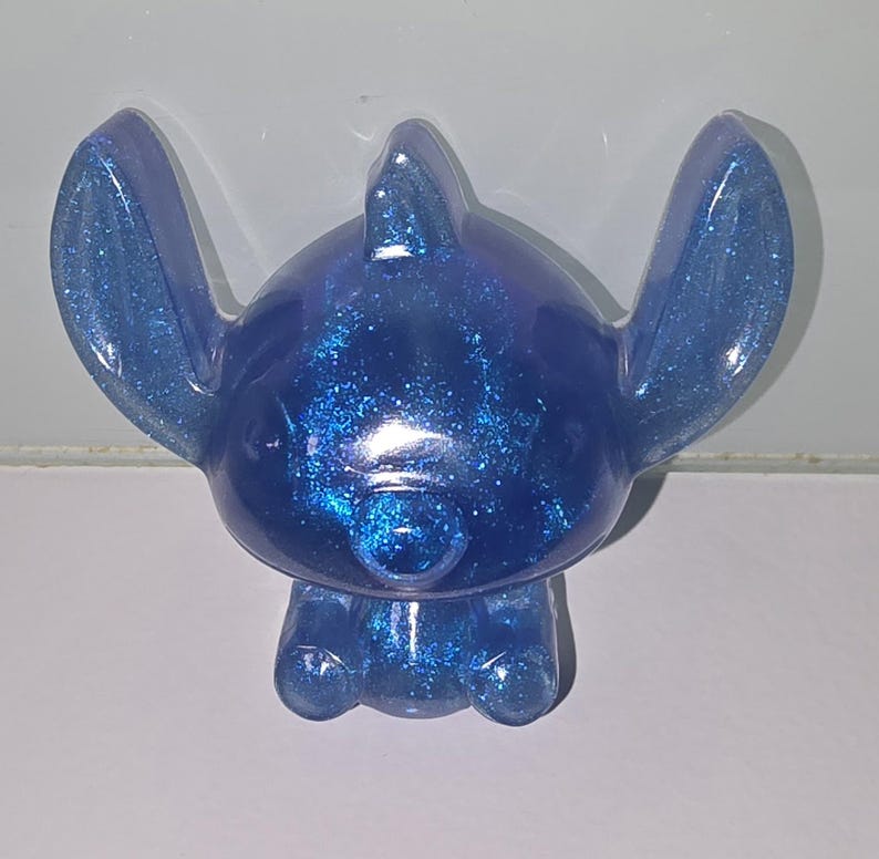 Op de afbeelding: Een blauw glitterfiguur van een stripfiguur met grote oren, een kleine neus en een afgerond lichaam. Het beeldje heeft een effen kleur met ingebedde glitter, waardoor het een sprankelend uiterlijk heeft. Het personage zit.