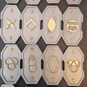 May include: A set of grey, shield-shaped metal tokens featuring gold and copper geometric designs. Each token displays a unique pattern, such as circles, diamonds, and angular shapes. The tokens are arranged in a grid formation.