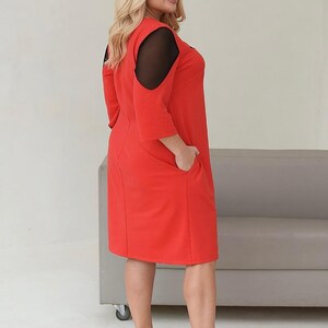 May include: A red dress with black mesh shoulder cutouts and three-quarter sleeves. The dress has pockets and is paired with black high heels. The model is wearing the dress and is standing in front of a gray couch.