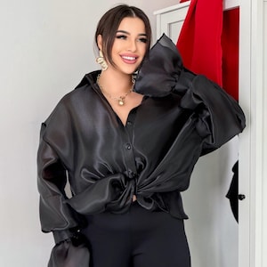 May include: Black satin button-down shirt with long sleeves and a knotted front. The shirt has a classic collar and cuffs with a ruffled design. The model is wearing black pants and carrying a black handbag. Gold jewelry completes the look.