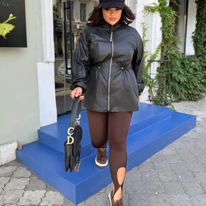 May include: A black hooded jacket with a front zipper and drawstring waist, paired with dark brown leggings featuring cut-out details at the ankles. The outfit includes a black baseball cap, brown lace-up sneakers, and a black handbag with gold hardware.