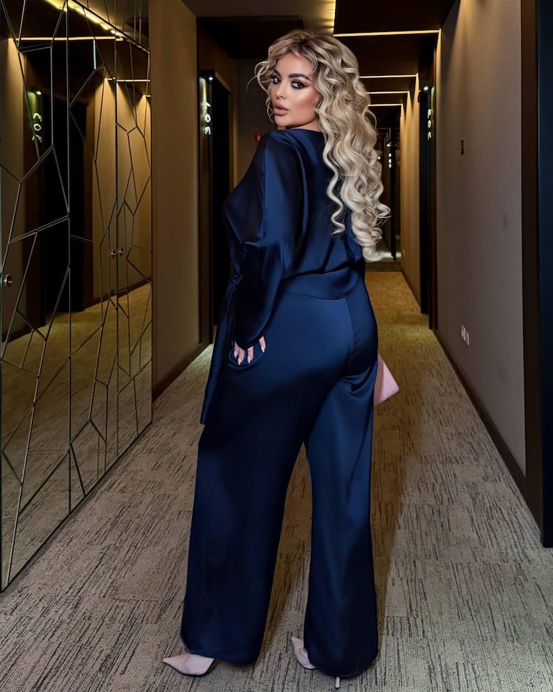 May include: A woman wearing a navy blue satin jumpsuit with wide-leg pants and a matching top. The top has long sleeves and a wrap-style design. The woman is standing in a hallway with a patterned carpet and a large mirror.