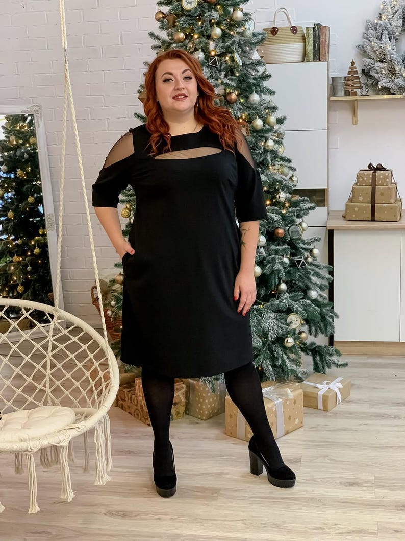 May include: A black dress with sheer shoulder and neckline accents. The dress has a knee-length hemline and short sleeves. The person is wearing black tights and platform heels. The background includes a Christmas tree with ornaments and wrapped gifts.