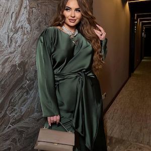 May include: An olive green satin dress with long sleeves and a wrap-style waist. The dress is paired with a small, taupe-colored handbag. The woman is wearing a necklace with a heart pendant.