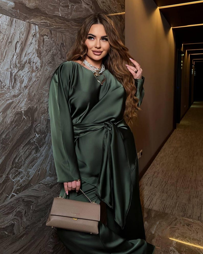 May include: An olive green satin dress with long sleeves and a wrap-style waist. The dress is paired with a small, taupe-colored handbag. The woman is wearing a necklace with a heart pendant.