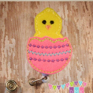 May include: A yellow chick peeking out of a pink egg with blue and purple embroidery. The egg is attached to a clear plastic badge holder with a gold clip.
