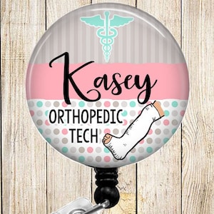 May include: A pink and gray round badge holder with a white medical symbol, the name "Kasey", and the text "Orthopedic Tech". The badge holder has a broken leg with a cast on it and a polka dot pattern.