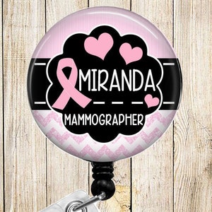 May include: A round, pink and black badge reel with the name "MIRANDA" and the title "MAMMOGRAPHER" in white text. The design includes a pink ribbon, hearts, and a chevron pattern. The reel has a clear plastic clip.