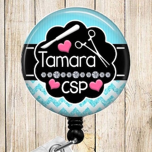 May include: A blue and black round badge holder with a white chevron pattern and pink hearts. The badge holder has the name "Tamara CSP" printed on it. The badge holder is attached to a clear plastic retractable badge reel.