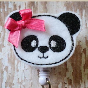 May include: A white felt panda with black eyes and ears, wearing a pink bow, attached to a retractable badge holder. The holder has a clear plastic badge holder and a silver clip.