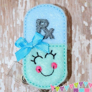 May include: A blue and green felt pill-shaped badge holder with a smiling face, eyelashes, and a pink bow. The top of the badge holder has the letter "R" embroidered in black.