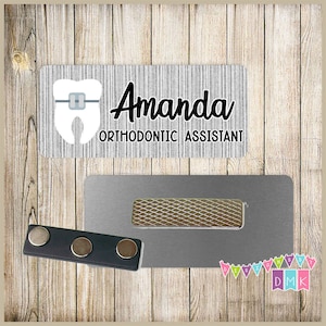 May include: A silver name tag with a gray and white tooth graphic and the text "Amanda Orthodontic Assistant". The name tag is attached to a silver magnetic holder with a black magnetic strip.
