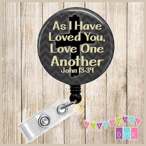 May include: Black and gray retractable badge holder with a round button featuring a black cross and the text "As I Have Loved You, Love One Another John 13:34".