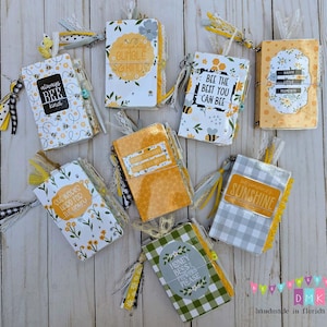 May include: A collection of small, decorative journals with bee-themed designs. The journals feature various patterns, including floral, gingham, and honeycomb, with phrases like "Always Bee Kind" and "Sunshine." Each journal has a tassel and ribbon.