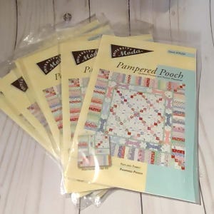 Może przedstawiać: Stos sześciu pakietów wzorów do patchworku z kolorowym wzorem patchworkowym. Pakiet z przodu przedstawia wzór patchworku o nazwie "Pampered Pooch" z pikowaną narzutą i podkładką pod garnek.