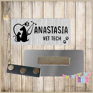 May include: A white rectangular name tag with black text and a paw print design. The text reads "Anastasia Vet Tech". The name tag is attached to a silver magnetic name tag holder.