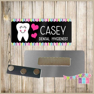 May include: A black and white name tag with a colorful striped border. The name tag features a cartoon tooth with pink hearts and the text "CASEY DENTAL HYGIENIST".